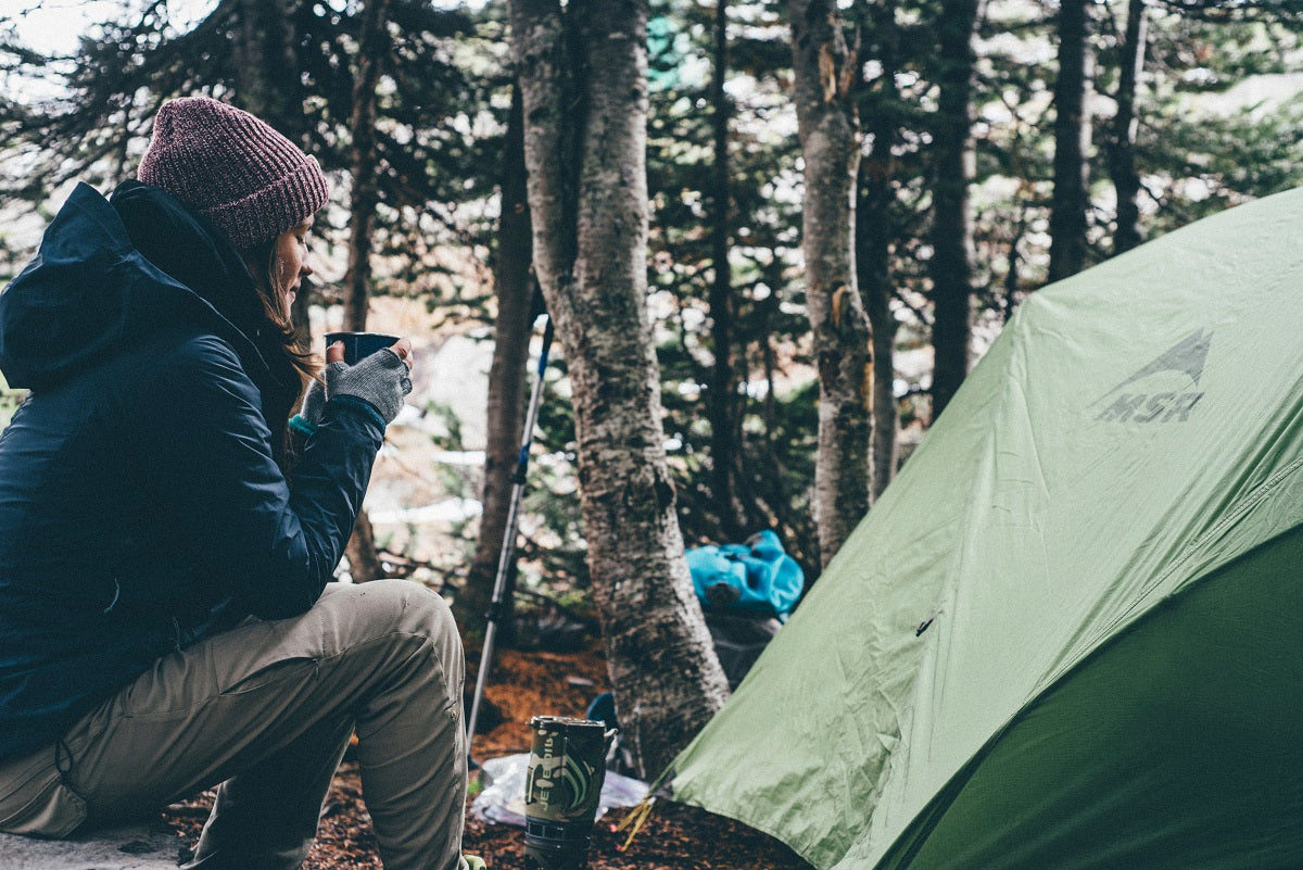 Woman beside fire pit and tent drinking coffee