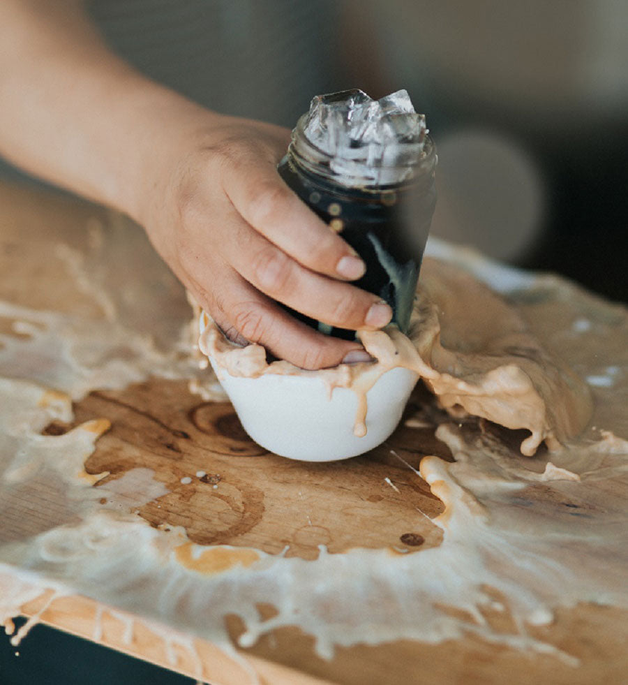 A hand places a glass of iced coffee into a mug containing a latte, coffee splashes everywhere