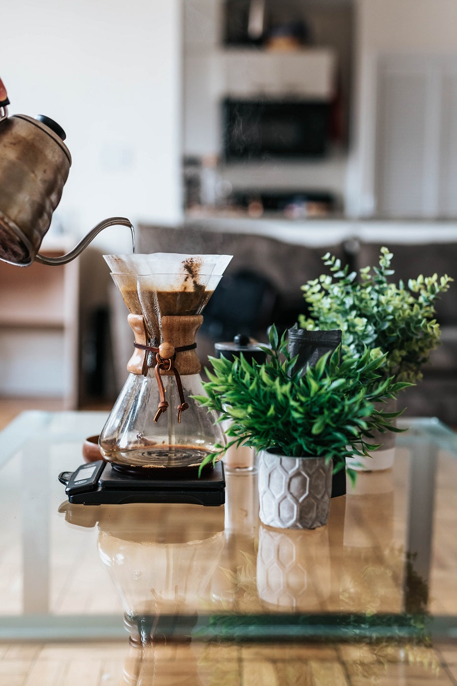 A Hand pours steaming water from a kettle into the top of a coffee loaded Chemex Pour Over brewer that sits on a modern glass coffee table