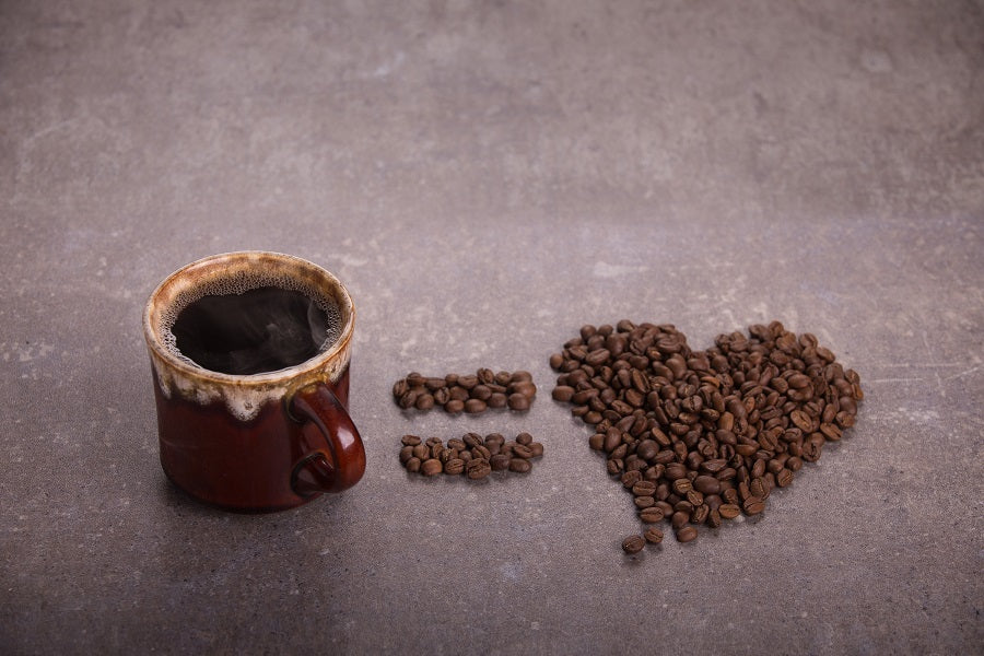 A cup of steaming coffee sits beside coffee beans which have been arranged into the shapes of an equals sign, and a heart