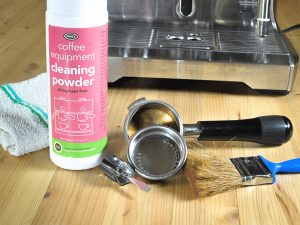 a bottle of coffee machine cleaning powder sits on a countertop alongside a flat-head screw driver and a paint-brush and assorted espresso machine parts