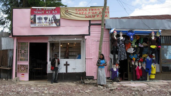 a street view of two shop fronts in Ethiopia, one is painted pink, the other blue, a woman stands out front wearing a blue top and grey skirt