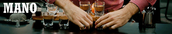 a short wide image of two hands carefully arranging a row of coffee-filled shot glasses on a countertop with the word Mano appearing in the center