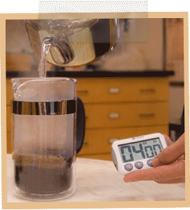 A hand holds a timer which reads 4 minutes, while hot water is poured over coffee grounds in a French Press